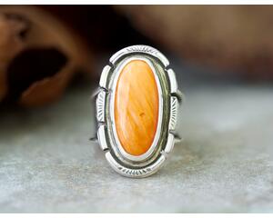 Oval orange spiny oyster cabochon in a bezel setting accented with stamped cutout wire on a wide silver band that is tapered and oxidized 6